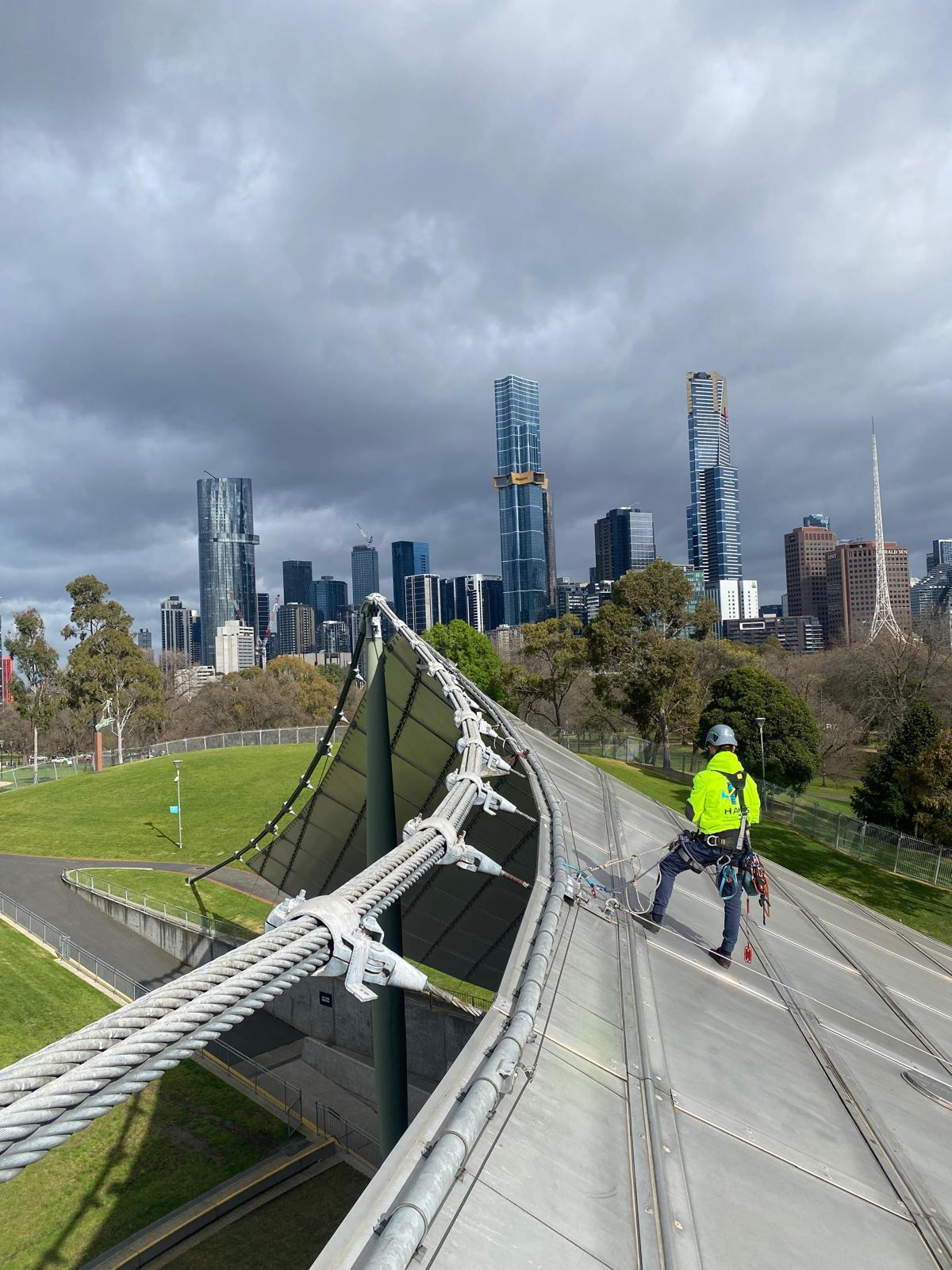 Rope access technician conducting high-rise maintenance on a stadium roof in Melbourne, secured with harness and IRATA-compliant gear, with city skyline in background.
